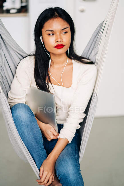 Продумана молода жінка з довгим темним волоссям тримає портативний ноутбук з навушниками, сидячи в гамаку в приміщенні — стокове фото