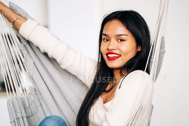 Young positive ethnic female sitting in hammock while laughing and resting at home looking at camera — Fotografia de Stock