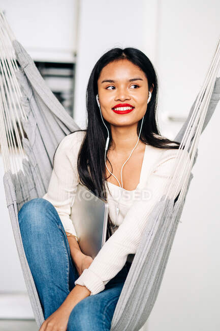 Smiling young woman with long dark hair holding a portable laptop with earphones while sitting in hammock indoors looking away — Photo de stock