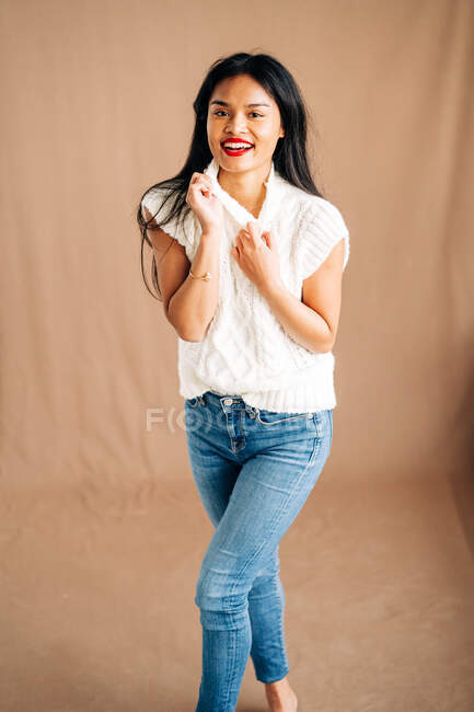 Satisfied ethnic female in casual outfit with toothy smile looking away while standing on brown background in studio — Photo de stock