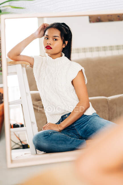 Отражение задумчивой этнической женщины в повседневной одежде с длинными волосами и яркими губами, смотрящей в зеркало — стоковое фото