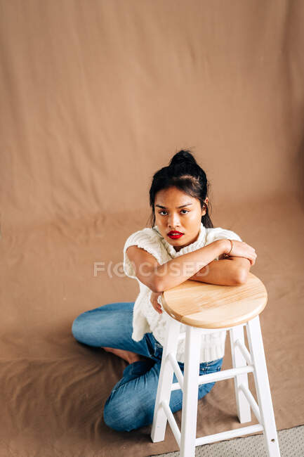 Cuerpo completo de joven hispana confiada usando ropa casual sentada en el piso apoyada en un taburete de madera en el estudio - foto de stock