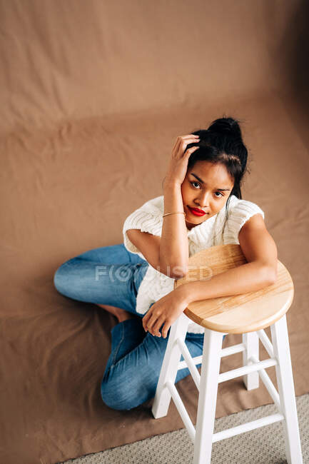 Full body of confident young Hispanic lady wearing casual clothes sitting on the floor leaning on wooden stool in studio — Photo de stock