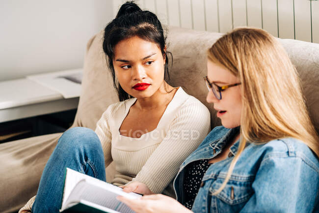 Glückliche Frau mit Laptop sitzt auf Sofa und kommuniziert mit nachdenklicher ethnischer Freundin — Stockfoto