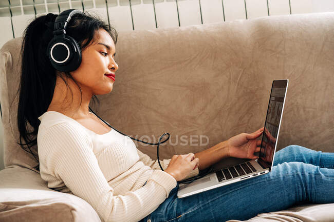 Seitenansicht einer fokussierten ethnischen Frau, die mit Kopfhörern auf einem Sofa liegt und an einem modernen Laptop arbeitet — Stockfoto