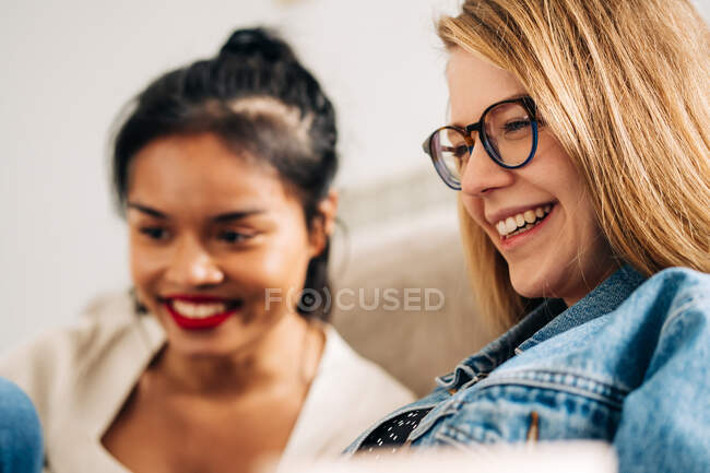 Happy woman with laptop sitting on sofa communicating with pensive ethnic female friend — Photo de stock