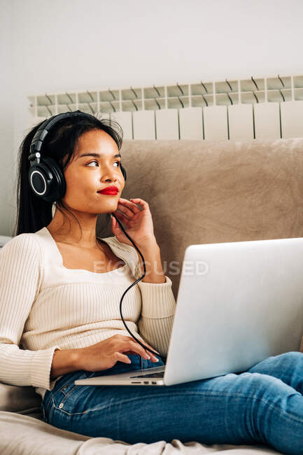 Seitenansicht einer fokussierten ethnischen Frau, die mit Kopfhörern auf einem Sofa liegt und an einem modernen Laptop arbeitet — Stockfoto
