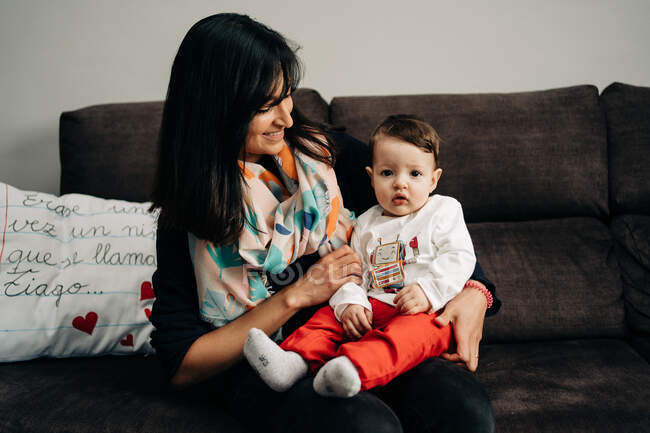 Happy ethnic woman smiling and holding cute little son on sofa at home — Fotografia de Stock