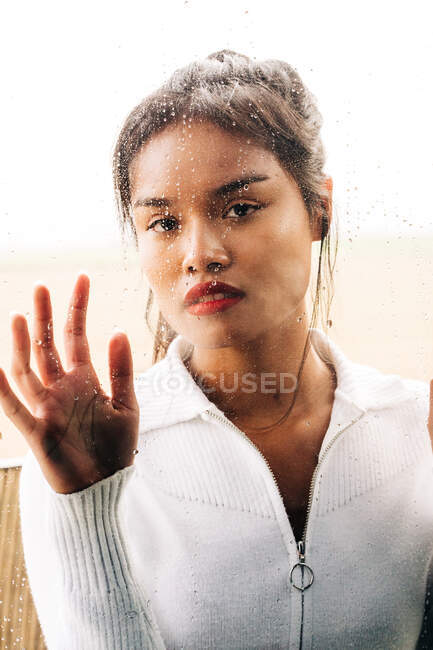Через стекло задумчивой этнической женщины с яркими губами, смотрящей в камеру при прикосновении капель — стоковое фото