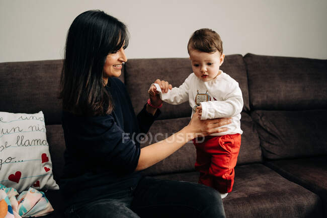Vista lateral de la feliz mujer étnica sonriendo y tomados de la mano de lindo hijo pequeño mientras juegan juntos en el sofá en casa - foto de stock