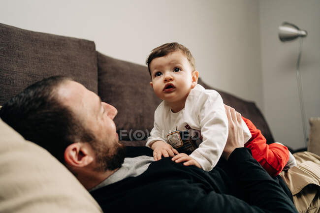 Low angle of positive young bearded father lying on comfortable couch and hugging adorable little son while spending time together at home — Photo de stock