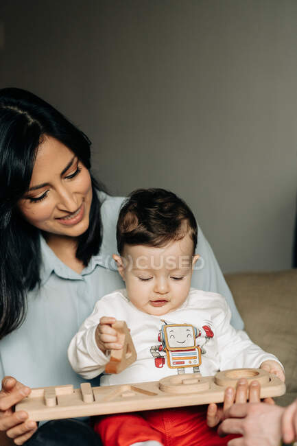 Crop young ethnic female with dark hair smiling while sitting on floor with cute little boy and playing with wooden toys at home — Photo de stock