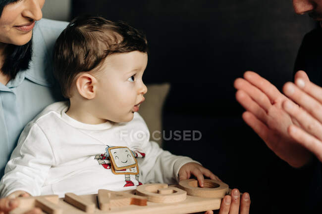 Ernte unkenntlich junger Vater umklammert Hände beim Spielen mit entzückenden kleinen Sohn sitzt auf Knien der ethnischen Mutter mit Holzspielzeug in den Händen — Stockfoto