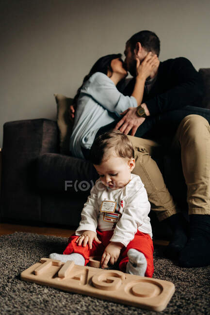 Giovani genitori irriconoscibili che si baciano sul divano vicino all'adorabile figlioletto che gioca sul pavimento con un giocattolo di legno con lettere di nome Tiago — Foto stock