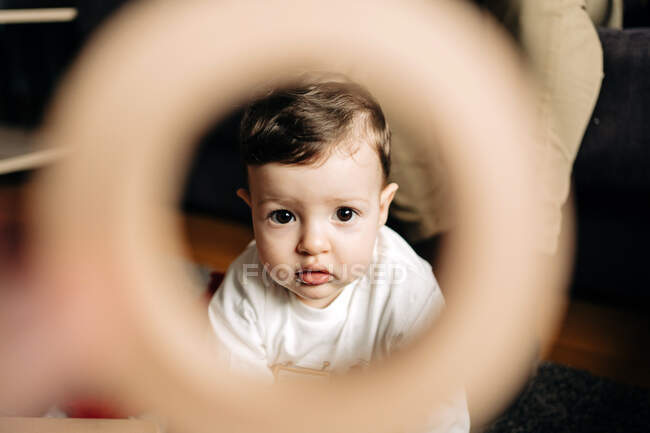 Through hole of adorable little boy sitting on floor and looking at camera at home — Photo de stock