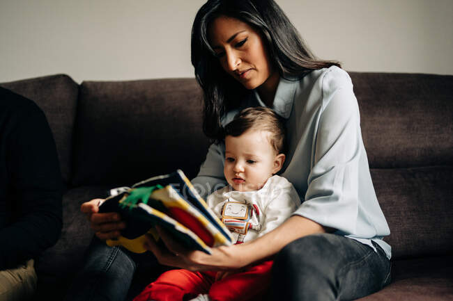 Crop young ethnic mother in casual clothes sitting on couch and reading book for adorable little son — Photo de stock