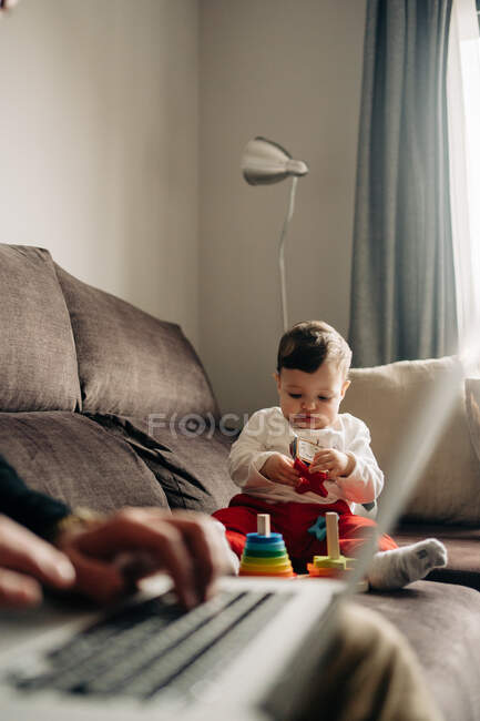Adorable little boy sitting on sofa and playing with colorful wooden cone tower near crop unrecognizable father typing on laptop during remote work at home — Photo de stock