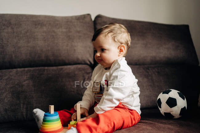 Side view adorable little boy sitting on sofa and playing with colorful wooden cone at home — Photo de stock
