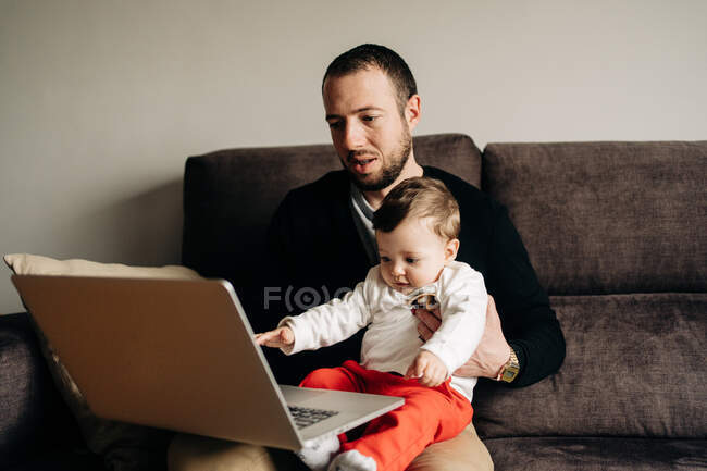 Young bearded man hugging little son while sitting on comfortable sofa and watching cartoon on laptop at home — Photo de stock