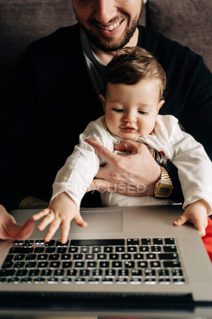 Обрізаний молодий бородатий чоловік обіймає маленького сина, сидячи на зручному дивані і дивлячись мультфільм на ноутбуці вдома — стокове фото