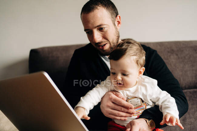 Молодий бородатий чоловік обіймає маленького сина сидячи на зручному дивані і дивиться мультфільм на ноутбук вдома — стокове фото