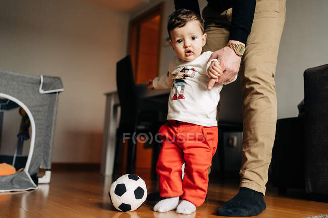 Little boy kicking ball while playing with cropped unrecognizable father at home — Photo de stock