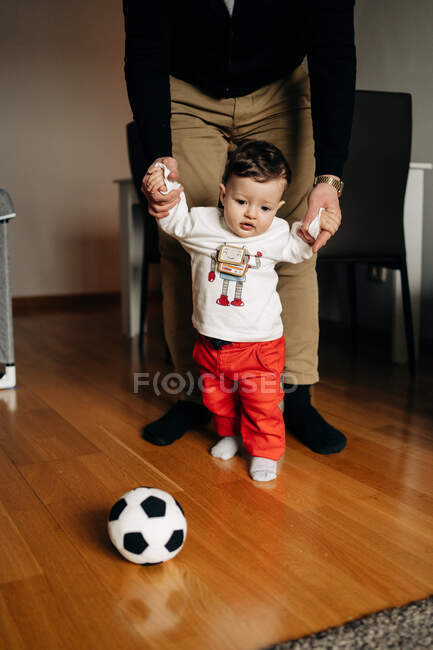 Little boy kicking ball while playing with cropped unrecognizable father at home — Fotografia de Stock