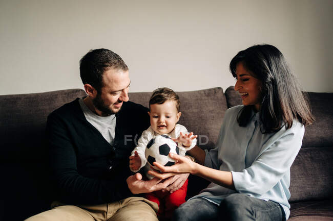 Amorosos padres jóvenes sentados en el sofá y abrazando a su pequeño hijo lindo jugando con la pelota en casa - foto de stock