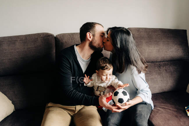 Loving young man and woman kissing each other while sitting on couch and and hugging cute little son playing with ball at home — Fotografia de Stock