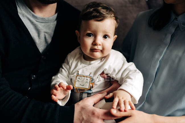 Crop unrecognizable young parents sitting on couch and embracing adorable little baby smiling and looking at camera — Photo de stock