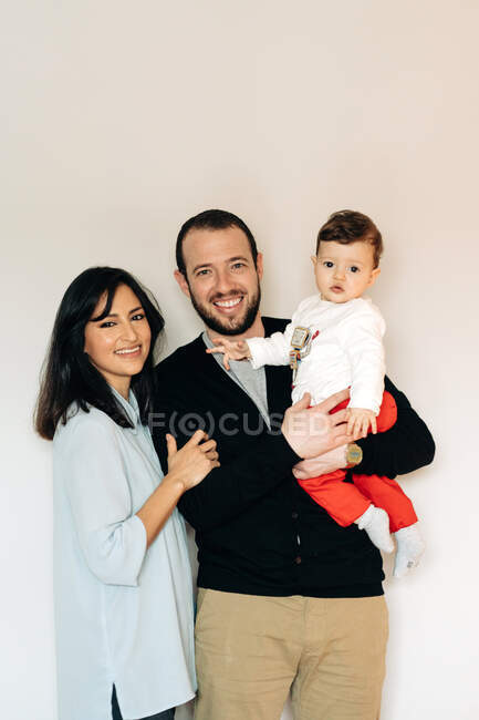Alegre joven multirracial madre y padre sonriendo mientras sostiene lindo feliz pequeño hijo contra fondo blanco mirando a la cámara - foto de stock