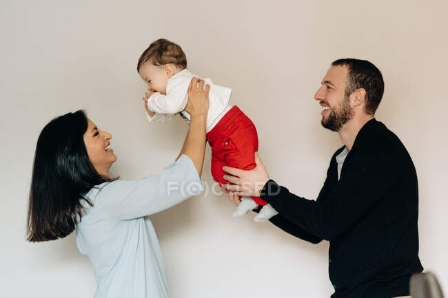 Vista lateral de la alegre joven madre multirracial y el padre sonriendo mientras abraza lindo hijo feliz contra el fondo blanco - foto de stock
