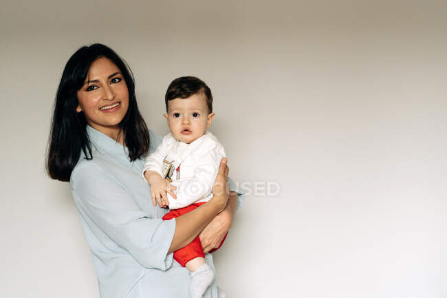Delighted young ethnic woman with long dark hair in casual clothes embracing cute little son and smiling against white wall — Stock Photo