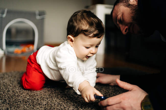 Батько в повсякденному одязі показує мультфільм на мобільному телефоні чарівному маленькому синові, сидячи разом на килимі у вітальні — стокове фото