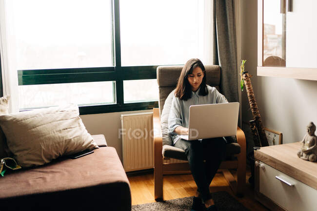 Концентрированная молодая этническая фрилансер с темными волосами в повседневной одежде сидит в удобном кресле и дистанционно работает на ноутбуке — стоковое фото