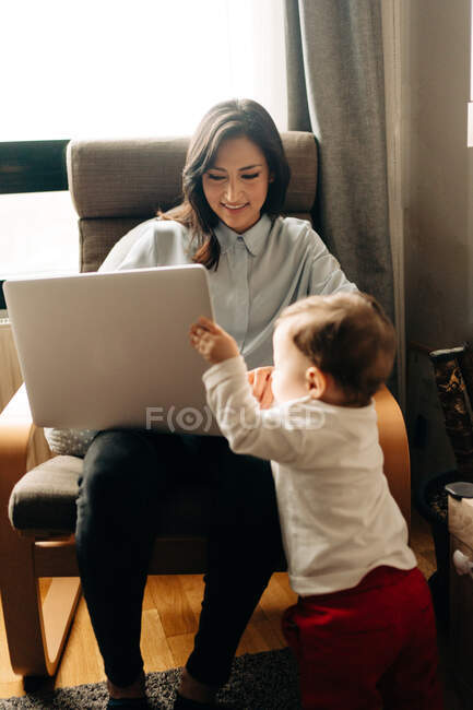 Zufriedene junge Mutter sitzt auf bequemem Stuhl und blättert im Netbook neben entzückendem kleinen Sohn — Stockfoto