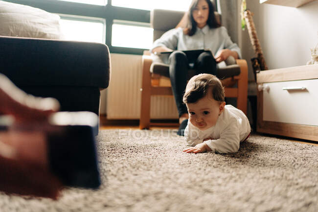 Lindo bebé arrastrándose en la alfombra hacia los padres mostrando video divertido en el teléfono móvil mientras pasan tiempo juntos en la sala de estar - foto de stock