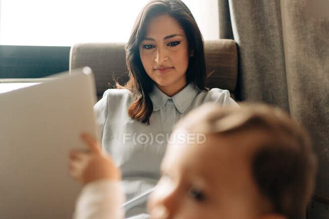 Contenuto giovane madre seduta su comoda sedia e navigazione netbook vicino adorabile piccolo figlio — Foto stock