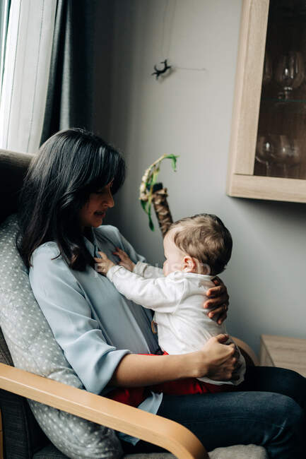Side view content young mother in casual wear sitting on cozy chair with adorable little baby on laps in modern living room — Fotografia de Stock