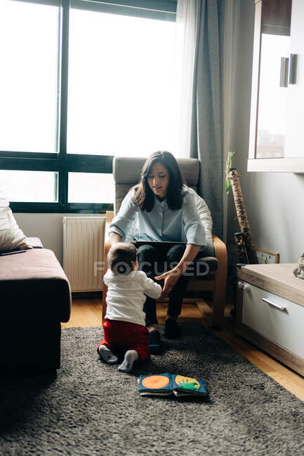 Зміст молода мати сидить на зручному стільці і переглядає нетбук біля чарівного маленького сина — стокове фото