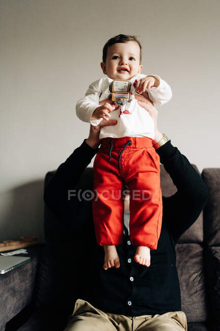Father sitting on comfortable couch and lifting cute smiling son on hands while spending time together in modern living room — Photo de stock