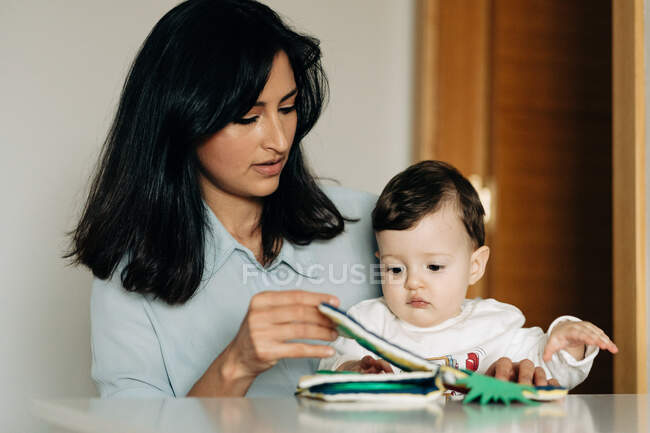 Positive young mother and adorable little son sitting at table and reading childrens book together — Photo de stock