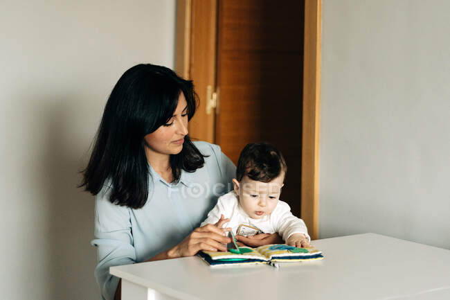 Positiva joven madre y adorable hijito sentado a la mesa y leyendo el libro de los niños juntos - foto de stock