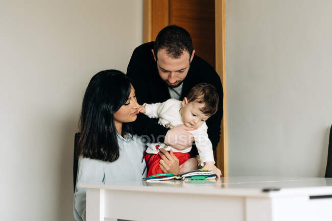 Positive young parents and adorable little son sitting at table and reading childrens book together — Fotografia de Stock