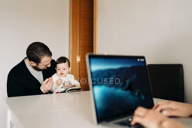 Young father and cute baby reading colorful childrens book while sitting at table near cropped unrecognizable mother working from home on netbook — Fotografia de Stock