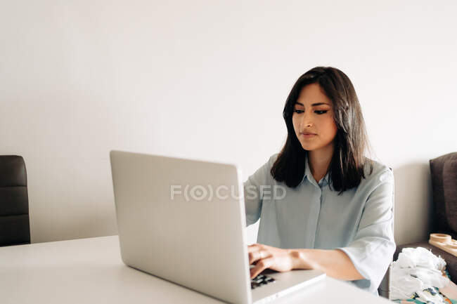 Mujer joven enfocada en blusa formal que navega por netbook moderno y se sienta a la mesa mientras trabaja remotamente en la sala de estar ligera - foto de stock