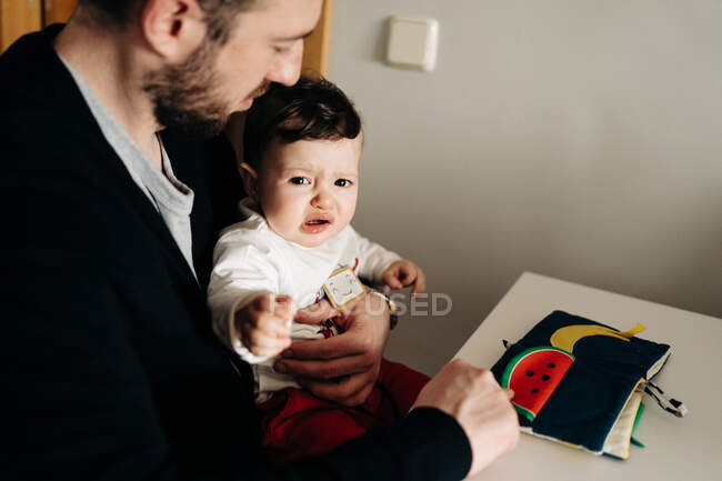 From above cropped positive young father and upset little son sitting at table and reading childrens book together — Fotografia de Stock