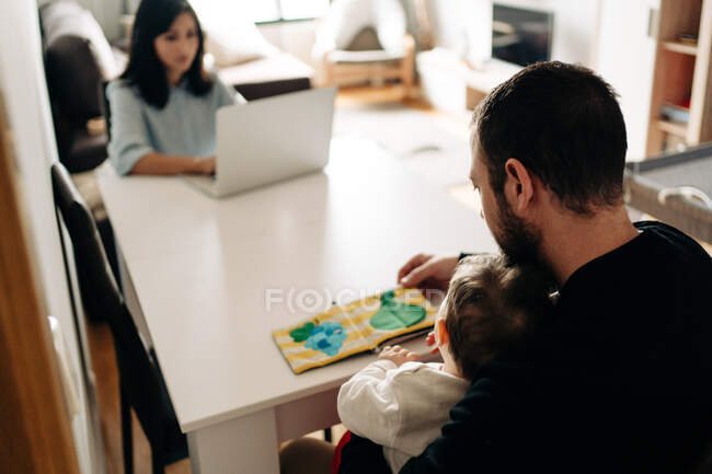 Молодой отец и симпатичный ребенок читают красочную детскую книгу, сидя за столом рядом с матерью, работающей из дома на нетбуке — стоковое фото