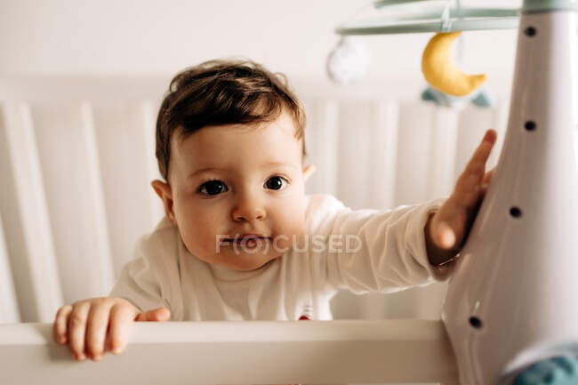 Сверху очаровательный маленький мальчик, стоящий в удобной кроватке и играющий с белой резиновой туфлей — стоковое фото