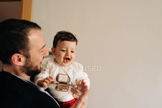 Cheerful young father holding adorable funny son on hands while having fun together in light room — Fotografia de Stock